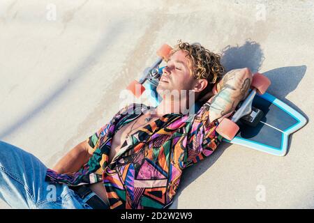 Alto angolo di giovane hippster tatuato maschio in elegante colorato camicia sdraiata sulla rampa di cemento con skateboard sotto la testa e. godendo l'estate nello skatepark Foto Stock