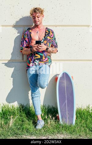 Maschio allegro hipster appoggiato sulla parete accanto allo skateboard prato nel parco e chiacchierare sui social media via cellulare telefono in giornata di sole Foto Stock