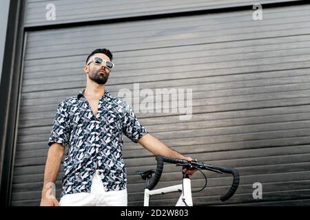 Da sotto serio bel maschio in abiti eleganti in piedi con bicicletta moderna vicino alle mura moderne della città mentre si guarda lontano Foto Stock