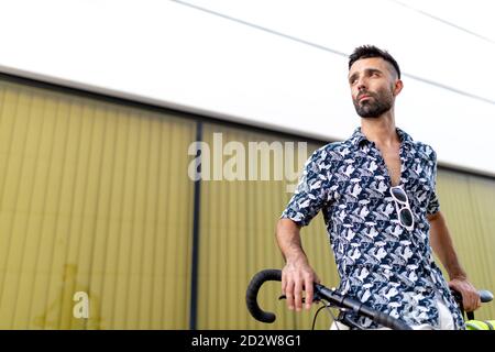 Da sotto serio bel maschio in abiti eleganti in piedi con bicicletta moderna vicino alle mura moderne della città mentre si guarda lontano Foto Stock