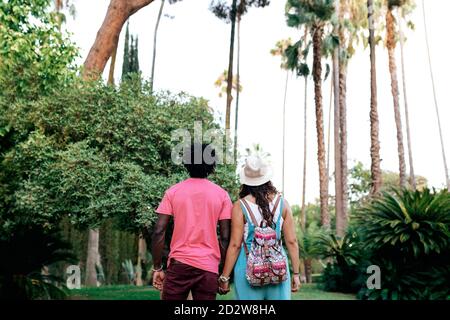 Vista posteriore di coppia multirazziale di viaggiatori in possesso di amore mani mentre si levano in piedi in un parco esotico e godendo la natura durante vacanze estive Foto Stock