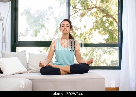 Seduta femminile flessibile concentrata con gambe incrociate e mani mudra Sul divano e facendo yoga in Padmasana mentre si pratica la consapevolezza con occhi chiusi Foto Stock