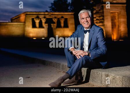 Positivo elegante senior maschile in elegante abito blu con prua cravatta e orologi da polso seduti al confine con la pietra nella città notturna e guardando via Foto Stock
