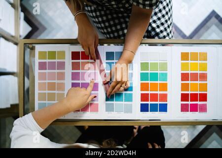 Vista dall'alto di designer femminili irriconoscibili in piedi al tavolo studio luminoso e scelta del colore dei campioni tessili Foto Stock