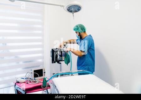 Vista laterale del medico di veterinario maschile in uniforme e maschera preparazione della macchina per anestesia per interventi chirurgici in sala operatoria luminosa in clinica veterinaria Foto Stock