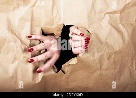 Foro apertura mani femmina in carta marrone. Rompere gli ostacoli. Fuga, protesta. Spazio di copia. Red nails.Woman's mani che raggiungono attraverso la carta strappata. Violen Foto Stock