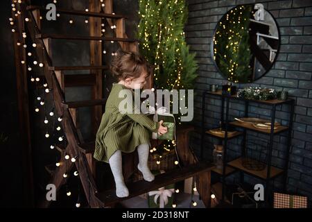 Una bambina in un vestito caldo verde si siede su una scala di legno, tenendo una scatola in un pacchetto con un arco. Regalo da Babbo Natale. Sfondo in luci, Natale Foto Stock