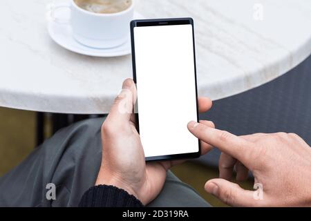 Primo piano dello smartphone portatile con le mani dell'uomo e schermata vuota dello spazio di copia. Immagine di un uomo seduto a un tavolo con un caffè in caffetteria e utilizzando mobil Foto Stock