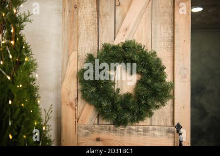 Una corona di Natale verde fatta di aghi di pino pende sulla porta eco di legno. Decorazioni per la casa festive, decorate con un ghirland con luci. A trama Foto Stock