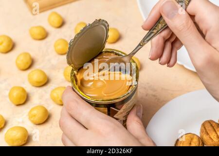 La mano della donna prende un cucchiaino da tè di latte condensato bollito da una lattina, accanto all'impasto di biscotti crudi e metà pronte per i biscotti noci Foto Stock