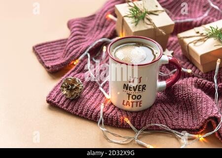 Una tazza di caffè con schiuma e regali di Natale su pullover in maglia viola su sfondo beige con ghirlanda. Primo piano Foto Stock