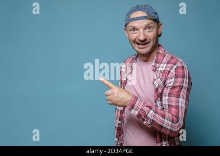 L'uomo divertente punta il dito indice a lato e guarda la telecamera con una strana espressione sul volto. Spazio di copia. Sfondo blu. Foto Stock