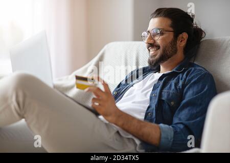 Guy allegro che usa il telefono mobile, la carta di credito ed il laptop che fornisce il pagamento in linea seduto sul lettino a casa. Foto Stock