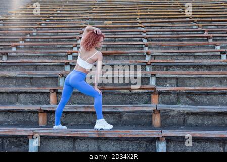 ritratto di una ragazza atleta dai capelli rossi, trentasei anni. su una panchina vuota di uno stadio di calcio, in posa, che mostra i muscoli del corpo. S Foto Stock