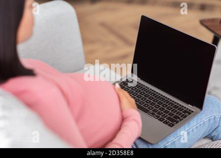 Donna incinta che usa un computer portatile con schermo vuoto seduto a casa Foto Stock