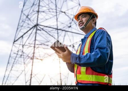 Immagine di ingegneria asiatica indossare abiti di sicurezza lavorando sulla torre ad alta tensione, controllare le informazioni sulla taplet. Foto Stock