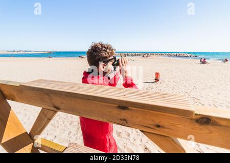 Un bagnino maschio caucasico che guarda attraverso il binocolo mentre indossa una maschera facciale, sulla spiaggia Foto Stock