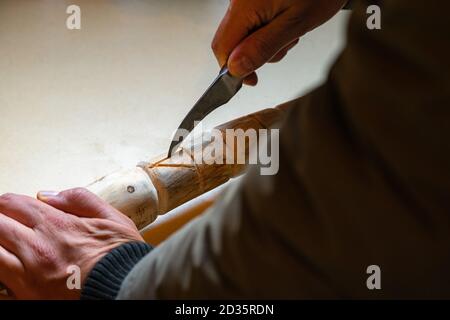 Processo di uomo che fa bastone di legno che cammina all'interno durante la quarantena. Bastone di legno intagliato sulla tavola usando il coltello Foto Stock
