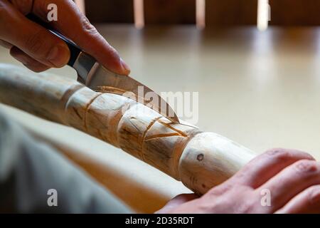 Processo di uomo che fa bastone di legno che cammina all'interno durante la quarantena. Bastone di legno intagliato sulla tavola usando il coltello Foto Stock