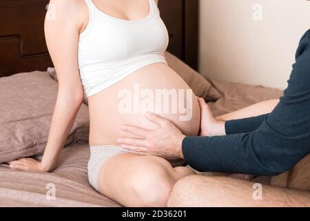 Donna incinta con marito seduto a letto, tenendo le mani sul ventre a casa. Foto Stock