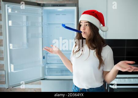 Giovane donna in cucina durante la quarantena. Persona femminile sconvolta in cappello rosso nel periodo di Natale o di nuovo anno. Frigorifero vuoto senza cibo o pasto, fruttato Foto Stock