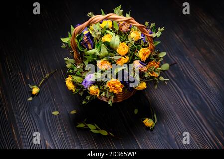 Fiori appassiti con foglie cadute su sfondo nero di legno. Cesto di vimini con rose secche gialle e blu. Arrangiamento dei fiori d'autunno. Bella d Foto Stock