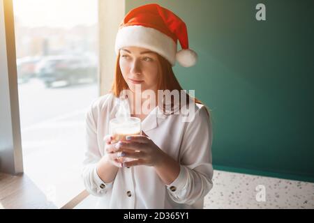 Il modello femminile in un cappello di natale tiene una tazza in mano. La giovane donna beve caffè e si guarda via. Foto Stock