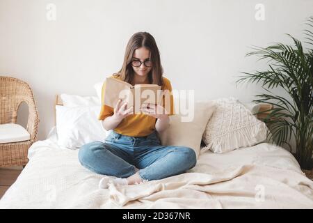 Giovane bella studentessa in occhiali, una T-shirt arancione e jeans blu si siede sul letto con cuscini e legge libro di carta. Concetto di auto-educazione all'hom Foto Stock