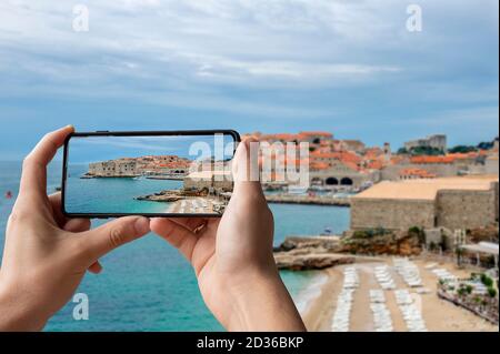 Turista scatta foto delle mura della fortezza di Dubrovnik, della spiaggia e delle acque turchesi. Famosa spiaggia di Banje con vista sulle mura della città di Dubrovnik e sulla città vecchia. Foto Stock