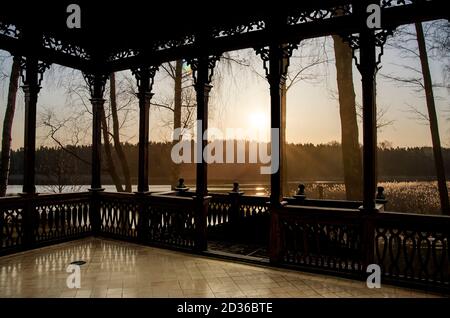 Vista dell'alba sul lago in primavera dalla galleria in legno o gazebo. Silhouette di foresta e i raggi del sole che sorge Foto Stock