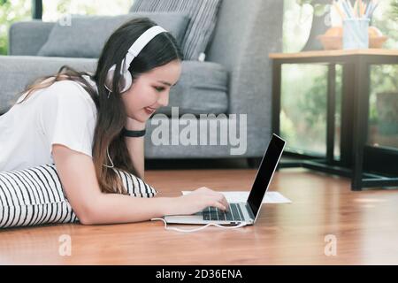 Una donna giovane e graziosa indossa le cuffie che si stendono sul pavimento e usano un computer portatile. Apprendimento a distanza. Luogo di lavoro remoto. Quarantena, coronovirus pandemia COV Foto Stock