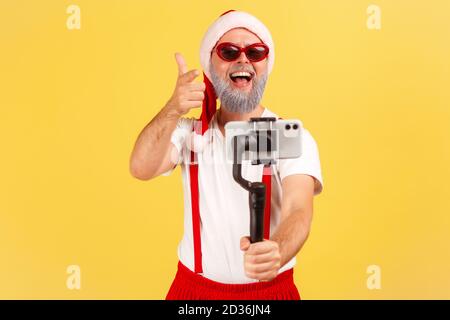 Felice positivo uomo anziano blogger in occhiali da sole e costume babbo natale che punta il dito alla videocamera di registrazione video o fare selfie su smartphone. IND Foto Stock