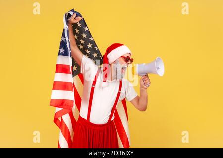 Uomo anziano serio in costume di babbo natale urlando a altoparlante che tiene bandiera degli stati uniti d'america, protesta patriottica sulle vacanze invernali. Poll Foto Stock