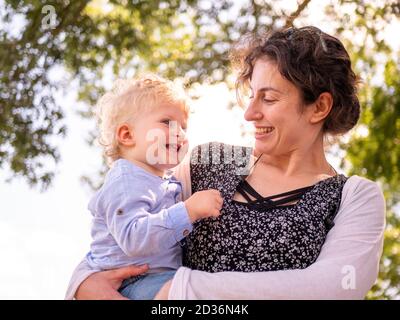 Madre orgogliosa e felice sorridente e mostrante il suo amore per il suo bambino di un anno ha tenuto in lei armi all'aperto Foto Stock