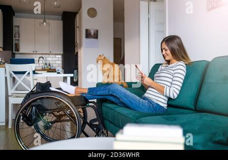 Giovane donna disabile che usa lo smartphone mentre si siede sul divano casa Foto Stock