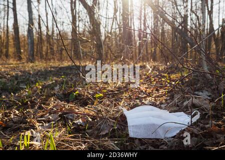 Maschera medica monouso bianca giace su vecchie foglie secche, gettato via come spazzatura nella foresta primaverile. Messa a fuoco selettiva Foto Stock