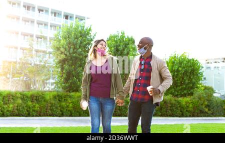 coppia interracial sorridente che indossa la maschera facciale che tiene un take away tazza di caffè mentre camminando in un parco in autunno.concetto circa la nuova normalità Foto Stock