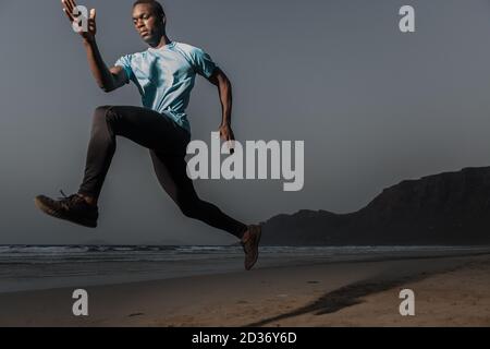 Da sotto sportivo nero serio che salta sulla sabbia vicino al mare durante l'allenamento fitness sulla spiaggia di Famara in serata a Lanzarote, Spagna Foto Stock