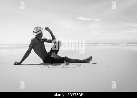Uomo nero giovane malgascio con il cappello e il grande sorriso seduto sulla spiaggia di sabbia bianca di fronte all'oceano in una posizione molto classica, appoggiato a mano Foto Stock