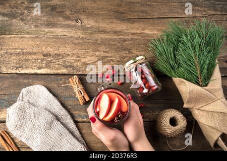 Le mani di una donna tengono un bicchiere di VIN brulé accanto ad un bouquet di rami di pino, mittene e bacche su sfondo ligneo. Foto Stock