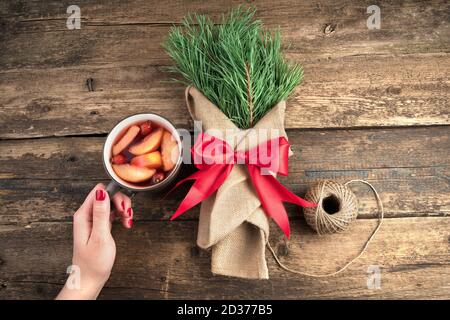 La mano di una donna tiene una tazza con una bevanda di frutta accanto ad un bouquet di rami di abete rosso e una palla di filo su uno sfondo naturale di legno. Foto Stock