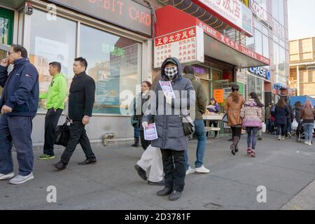 Una donna cinese americana in una maschera passa fuori volantini pubblicitari su Main St. A Chinatown, Flushing, New York. Nel 2016, prima della pandemia. Foto Stock