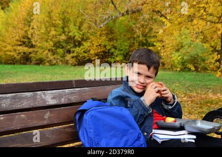 Primo piano di un piccolo scolaro seduto su una panchina del Parco e aprendo lo zaino della scuola durante una pausa pranzo. Mangiare un panino fuori da una scatola per il pranzo. Foto Stock