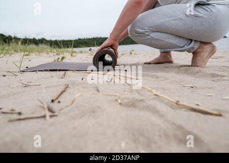 L'uomo dispiega un tappeto da yoga su una spiaggia sabbiosa vicino a un lago, sta per fare esercizi all'aperto in una giornata estiva. Foto Stock