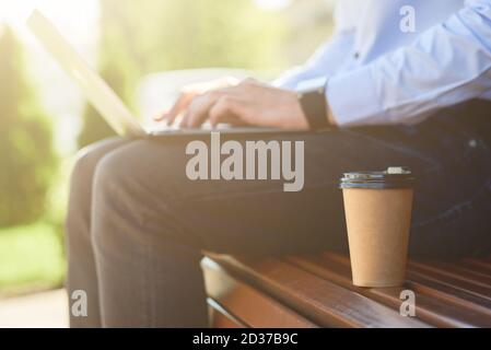 Un'immagine ritagliata di un freelance maschile che lavora online su un computer portatile e beve caffè mentre si siede sulla panchina all'aperto in una giornata di sole, vista laterale Foto Stock
