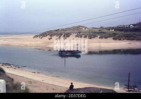 Una barca da pesca torna sul fiume fino al porto di Hayle in Cornovaglia, guardata da un ragazzo seduto su una scatola della pillola della seconda guerra mondiale. Photo taken in 1960's , on Agfa CT18 Foto Stock