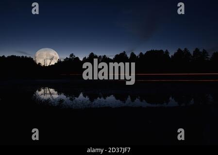 Riflessione di sentieri di luce da un veicolo su un piccolo lago di notte, contro gli alberi e il cielo notturno. Foto Stock