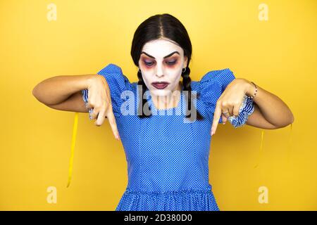 donna che indossa un costume di halloween spaventoso bambola su sfondo giallo sorpreso, guardando la fotocamera e puntando con le dita e le braccia sollevate Foto Stock