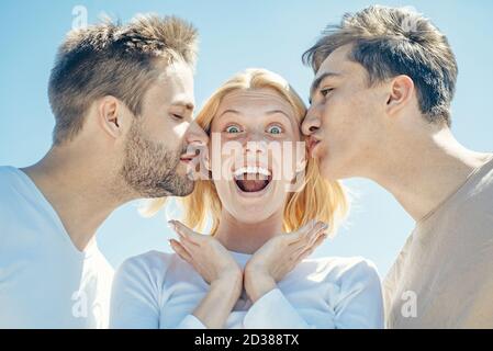 Bionda ragazza felice ottenere baci da due bei ragazzi. Donna affascinante in piedi tra due amici. Il concetto dei migliori amici. Gruppo di amici in chiaro Foto Stock