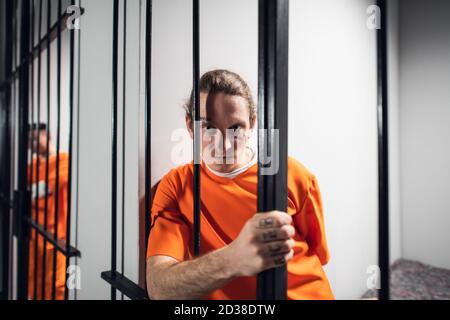 Un maniaco criminale disperato con tatuaggi in una cella solitaria è tenuto dietro le barre. Un'occhiata dura alla fotocamera Foto Stock